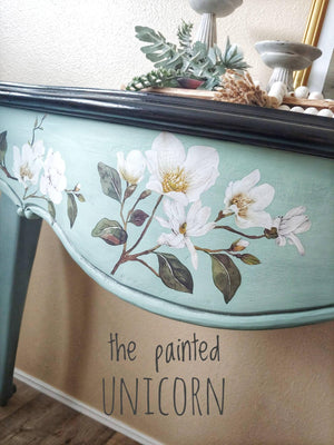 May include: A close-up of a painted, light blue table with a black trim. The table is decorated with white magnolia flowers and green leaves. The text "the painted UNICORN" is visible on the table.